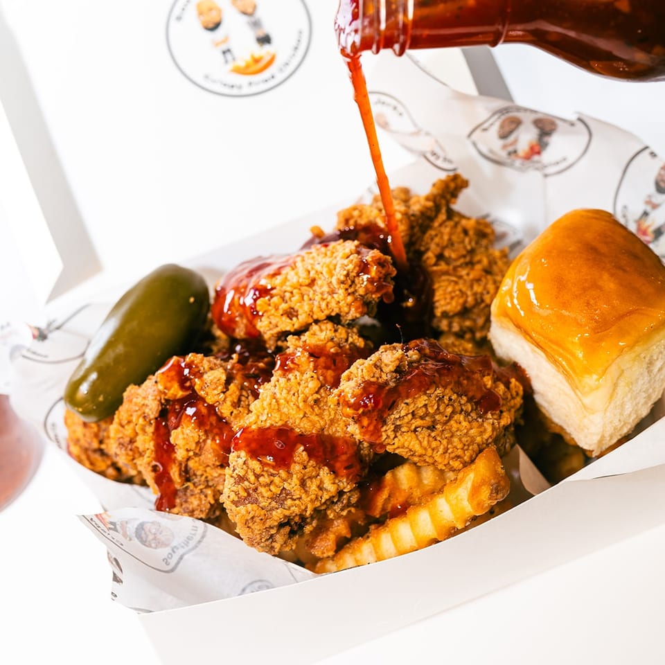 Crispy fried chicken pieces drizzled with red sauce, crinkle-cut fries, pickles, and a cheeseburger in a takeaway box.