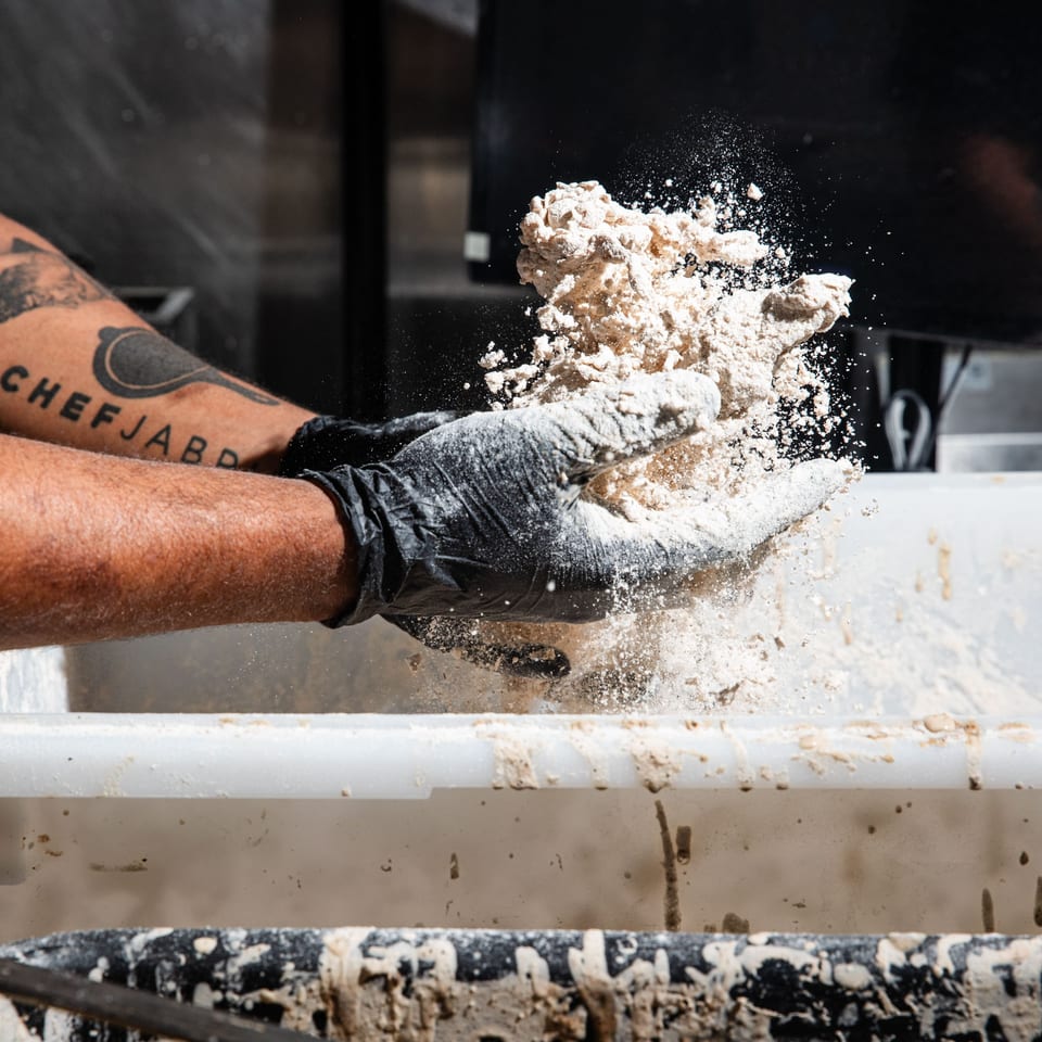 Gloved hands shape a mound of flour-dusted dough while flour scatters across a bakery work surface.