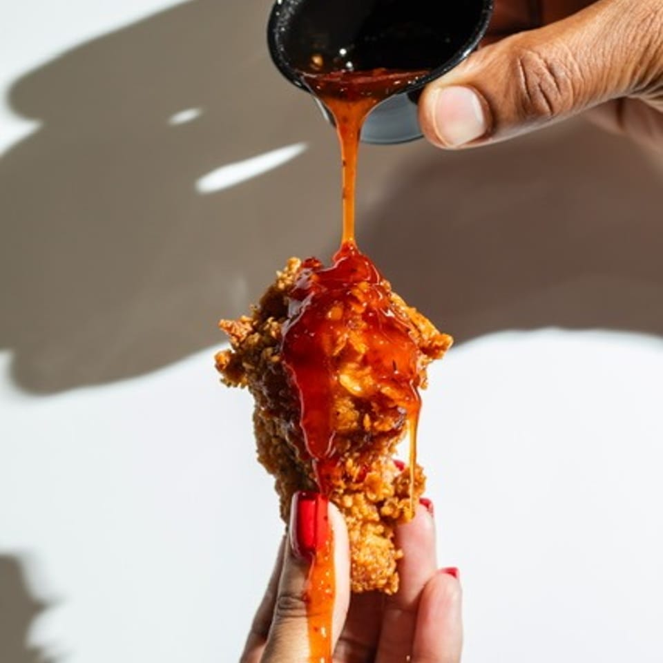 Hands drizzle glossy caramel sauce over a crispy fried dessert bar against a bright white background.
