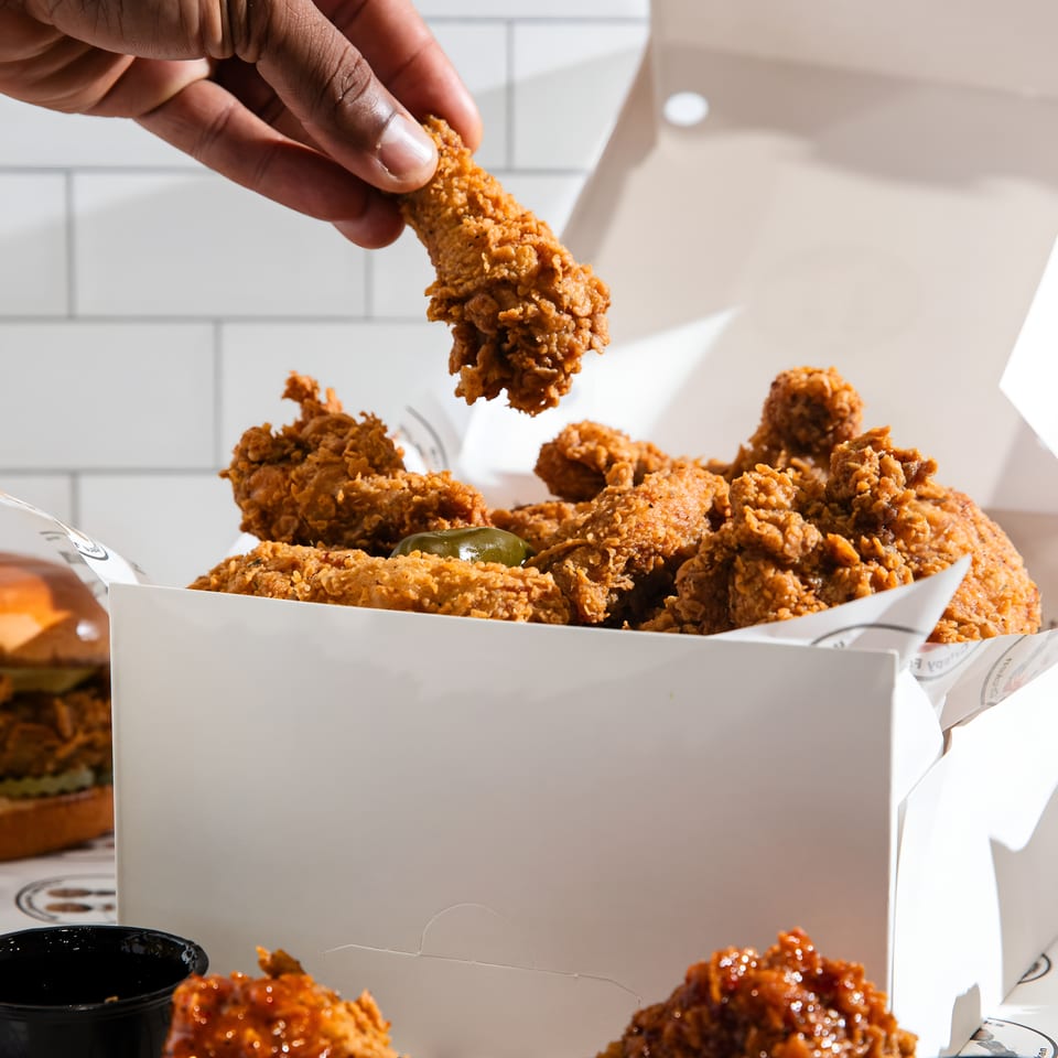 Hand places crispy fried chicken into a takeout box filled with chicken pieces, dipping sauces, and a green pepper.
