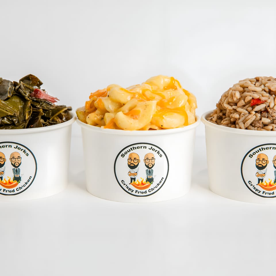 Three Southern Jerks containers hold leafy greens, creamy macaroni and cheese, and seasoned rice against a white background.