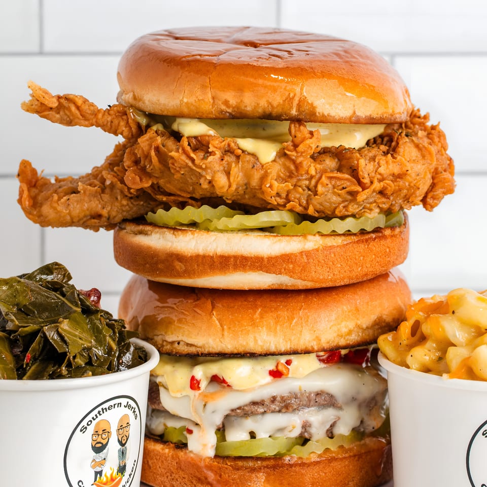 Stacked fried chicken and cheeseburger sandwiches with pickles, creamy sauce, collard greens, macaroni and cheese, and branded Southern Jerks cups.