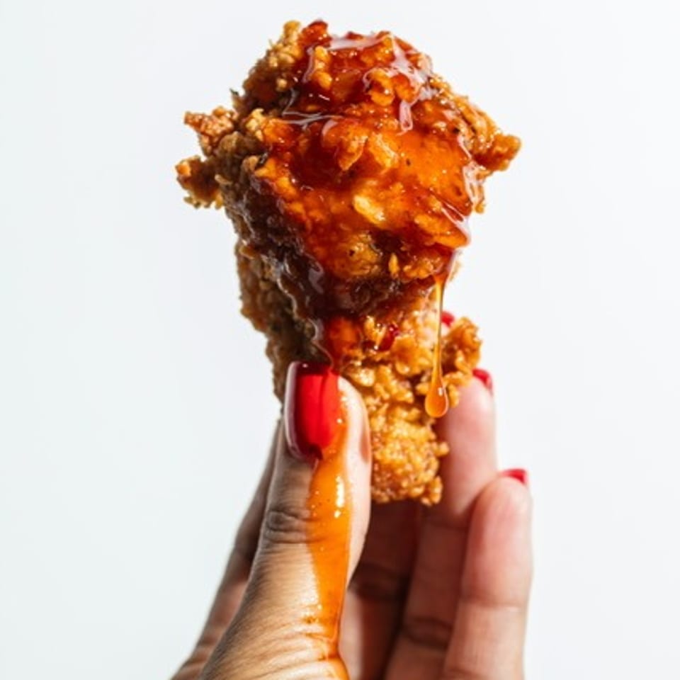 Hand with red-painted nails holding a crispy fried chicken drumstick glazed in glossy orange sauce.