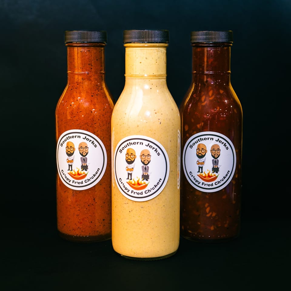 Three bottles of Southern Jerks crispy fried chicken sauce in red, creamy, and dark varieties against a black background.